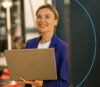 A smiling businesswoman in a blue blazer is holding a laptop